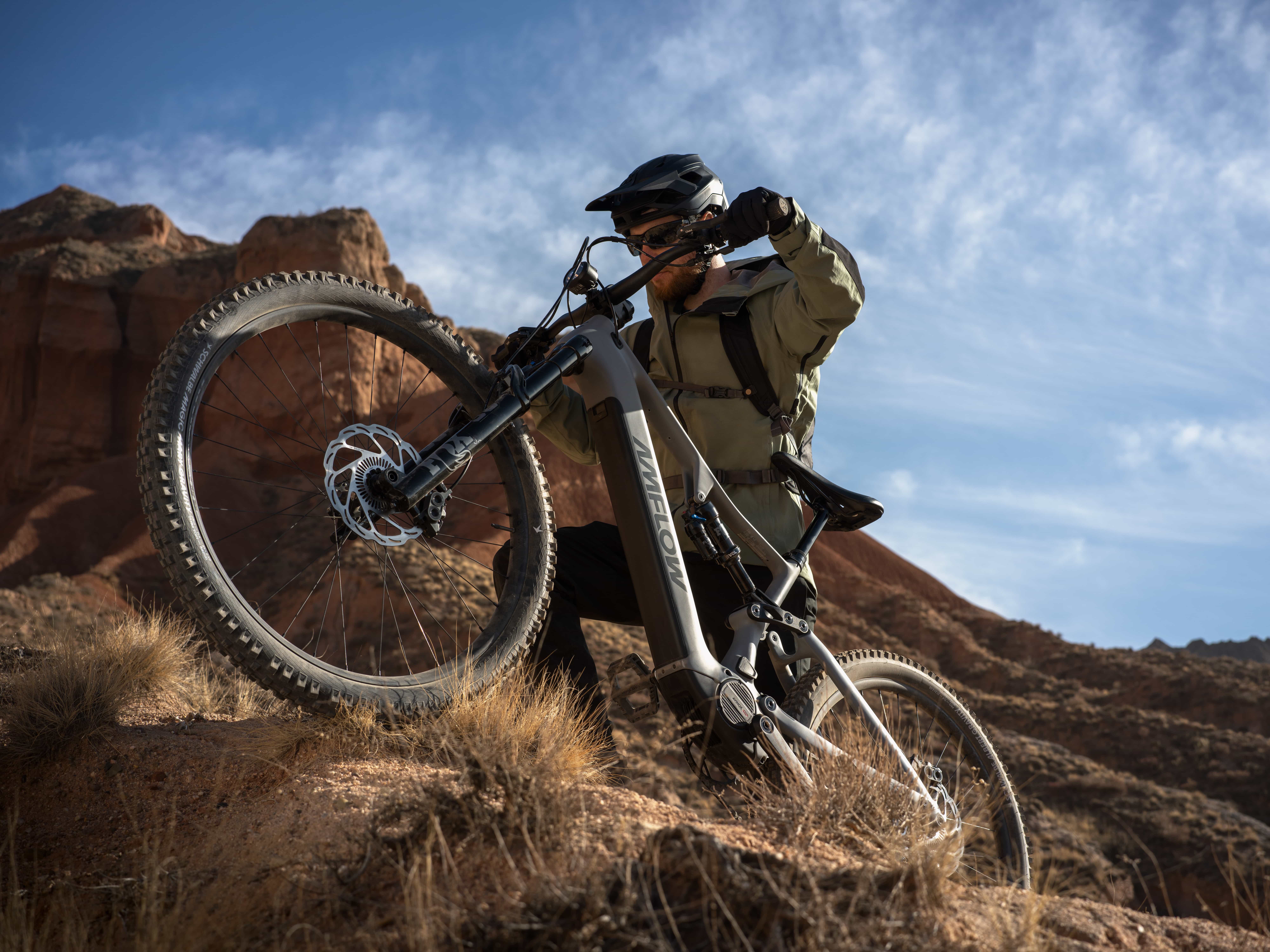 Cyclist pushing a silver AMFLOW full-suspension electric mountain bike up a rocky red-rock desert trail